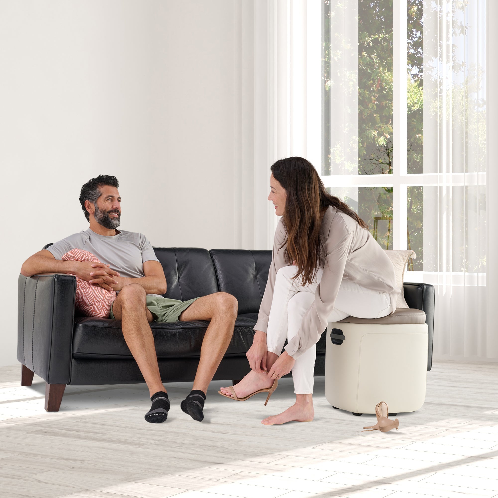Reveal Ottoman Massager shown in bright living room with person relaxing on couch while partner demonstrates foot massage feature